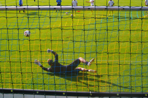 Ein Fussballspieler hält den Ball, vor einem Tor mit Netz im Vordergrund und einigen Menschen im Hintergrund, sowie mehreren Bällen auf dem Boden verteilt.