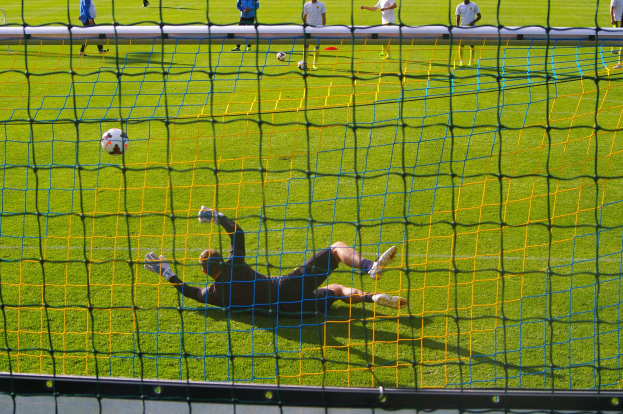 Ein Fussballspieler hält den Ball, vor einem Tor mit Netz im Vordergrund und einigen Menschen im Hintergrund, sowie mehreren Bällen auf dem Boden verteilt.