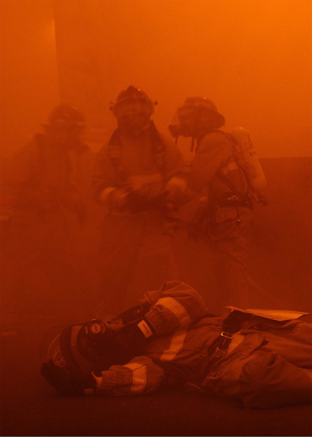 Schwarz-weißes Bild eines Feuerwehrmanns, der vor intensiven Flammen auf dem Boden liegt, umgeben von einer Gruppe von Helden mit Sauerstoffflaschen.
