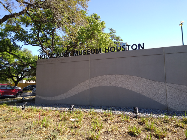 Das Holocaust-Museum in Houston, Texas, mit einer textbedeckten Wand, Steinen, Gras, Pflanzen, einer Baumgruppe, Fahrzeugen auf der Straße, einem Straßenpfahl und einem bewölkten Himmel.