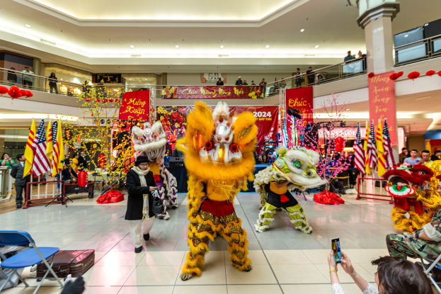 Ein lebendiger chinesischer Neujahrsfeier in einem Einkaufszentrum mit tanzenden Menschen in Kostümen und anderen mit Mobiltelefonen sitzend, umgeben von Flaggen, Bannern, Dekorationen und Lichtern.