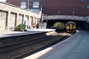 Ein Zug fährt auf den Schienen mit Plattformen auf beiden Seiten, einer Hauswand links, Straßenlaternen, Pflanzen, Bänken, einer Hütte, einer Brücke über die Plattformen und Gebäuden und Masten im Hintergrund.