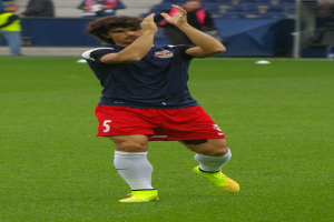 Ein Mann in einem blauen Hemd und roten Shorts steht auf einem Fußballfeld bereit zum Spielen, mit einer Gruppe von Menschen im Hintergrund.