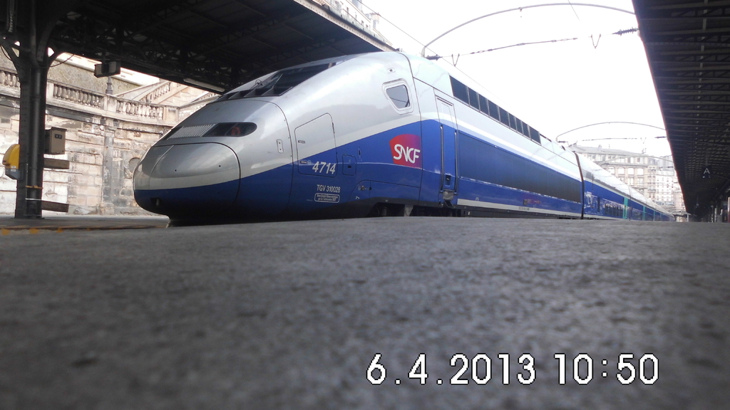 Ein Zug ist in der Mitte des Bildes mit einem Bahnsteig und Text unten, ein Gebäude mit Dach und Säule links und Gebäude mit Dächern, Kabeln und Himmel oben.