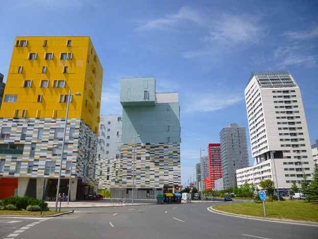Eine Stadtstraße mit hohen Universitätsgebäuden im Hintergrund, Straßenlaternen, Fahrzeugen, Infotafeln, Grünflächen und einem bewölkten Himmel.