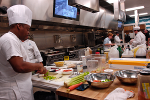 Eine Gruppe von Köchen in einer Küche, mit einem im Vordergrund in einer weißen Schürze und Mütze, der ein Messer hält und von verschiedenen Utensilien, Schüsseln, Gemüse, einem Handy und Flaschen umgeben ist, mit einem Fernseher und Deckenlampen im Hintergrund.