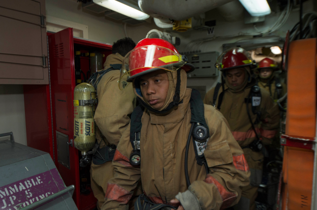 Eine Gruppe von Feuerwehrleuten in Uniformen und Helmen, einer hält einen Feuerlöscher, mit einem Metallobjekt links und Lampen, Rohren und anderen Gegenständen im Hintergrund.