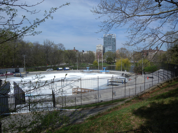 Eisstadion umgeben von einem Metallzaun, Gras, Bäumen, Fahrzeugen, Straßenlaternen, einem Gewässer, Gebäuden und einem bewölkten Himmel in einer Parkanlage.