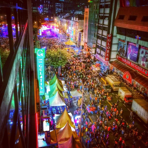 Eine lebhafte Karnevalsszene mit einer großen Menge auf der Straße, umgeben von Zelten, Geschäften, Gebäuden und Bannern, mit zahlreichen Lichtern im Hintergrund.