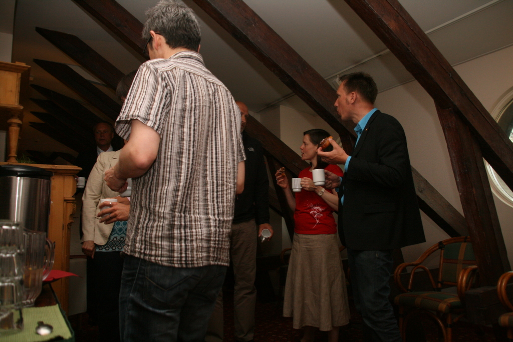 Eine Gruppe von Menschen mit Tassen in der Hand, links ein Becher und ein Tisch, oben ein Dach und rechts Stühle.