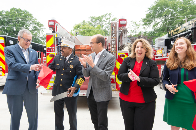 Eine Gruppe von Menschen steht vor einem Feuerwehrauto, klatscht und lächelt, wobei zwei eine Schere und ein rotes Tuch halten, während einer Zeremonie zur Einweihung der Feuerwache Alexandria, mit Bäumen, Kabeln und einem klaren blauen Himmel im Hintergrund.