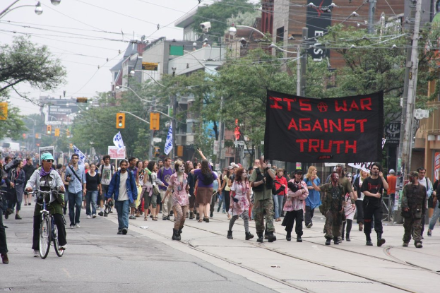 Eine Gruppe von Menschen marschiert mit Fahnen und Schildern die Straße entlang, daneben ein Radfahrer, Laternenpfähle, Stromkabel, Ampeln, Schilder, Bäume und Gebäude im Hintergrund.