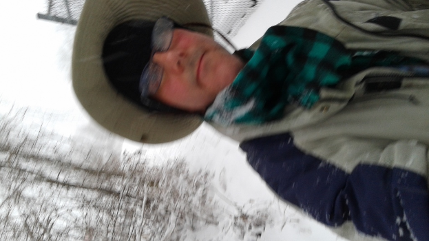 Eine Person in Jacke und Hut vor einem verschneiten Hintergrund mit Bäumen links unten.