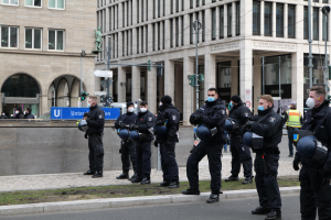 Eine Gruppe von Polizisten in schwarzen Uniformen und Masken steht vor einem Glasfenstergebäude mit Säulen in Berlin, Deutschland, einige halten Helme, mit Laternen, Verkehrszeichen, Texttafeln, einer Statue, anderen Menschen und einem grasbewachsenen Boden im Hintergrund.