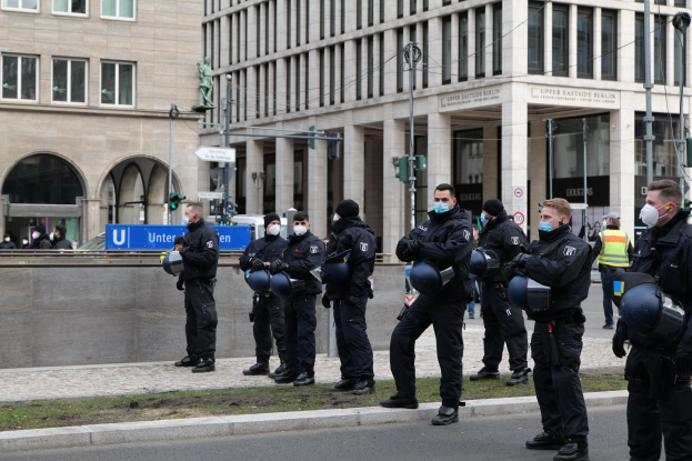 Eine Gruppe von Polizisten in schwarzen Uniformen und Masken steht vor einem Glasfenstergebäude mit Säulen in Berlin, Deutschland, einige halten Helme, mit Laternen, Verkehrszeichen, Texttafeln, einer Statue, anderen Menschen und einem grasbewachsenen Boden im Hintergrund.