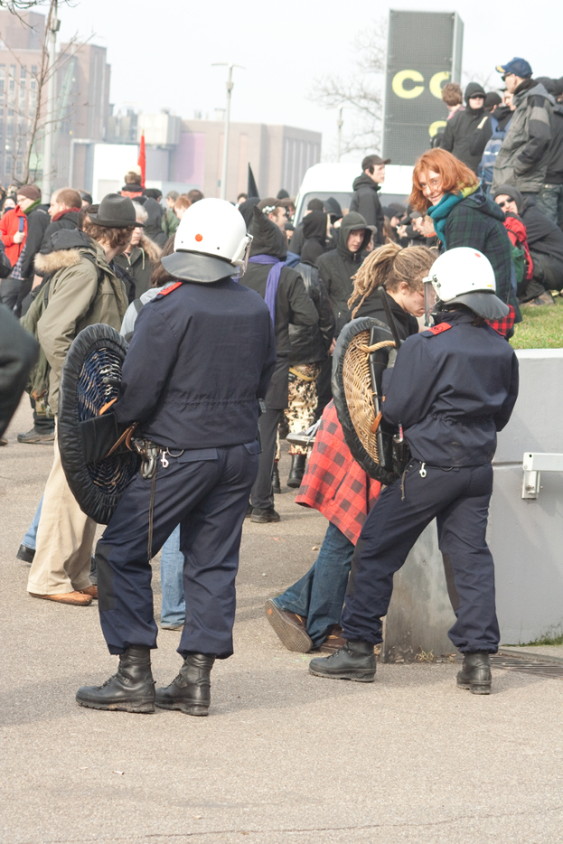 Eine Gruppe von Menschen, die auf einer Straße gehen, mit zwei Personen in der Mitte, die wie Polizeibeamte aussehen, Gebäuden im Hintergrund und Boden unten im Bild.