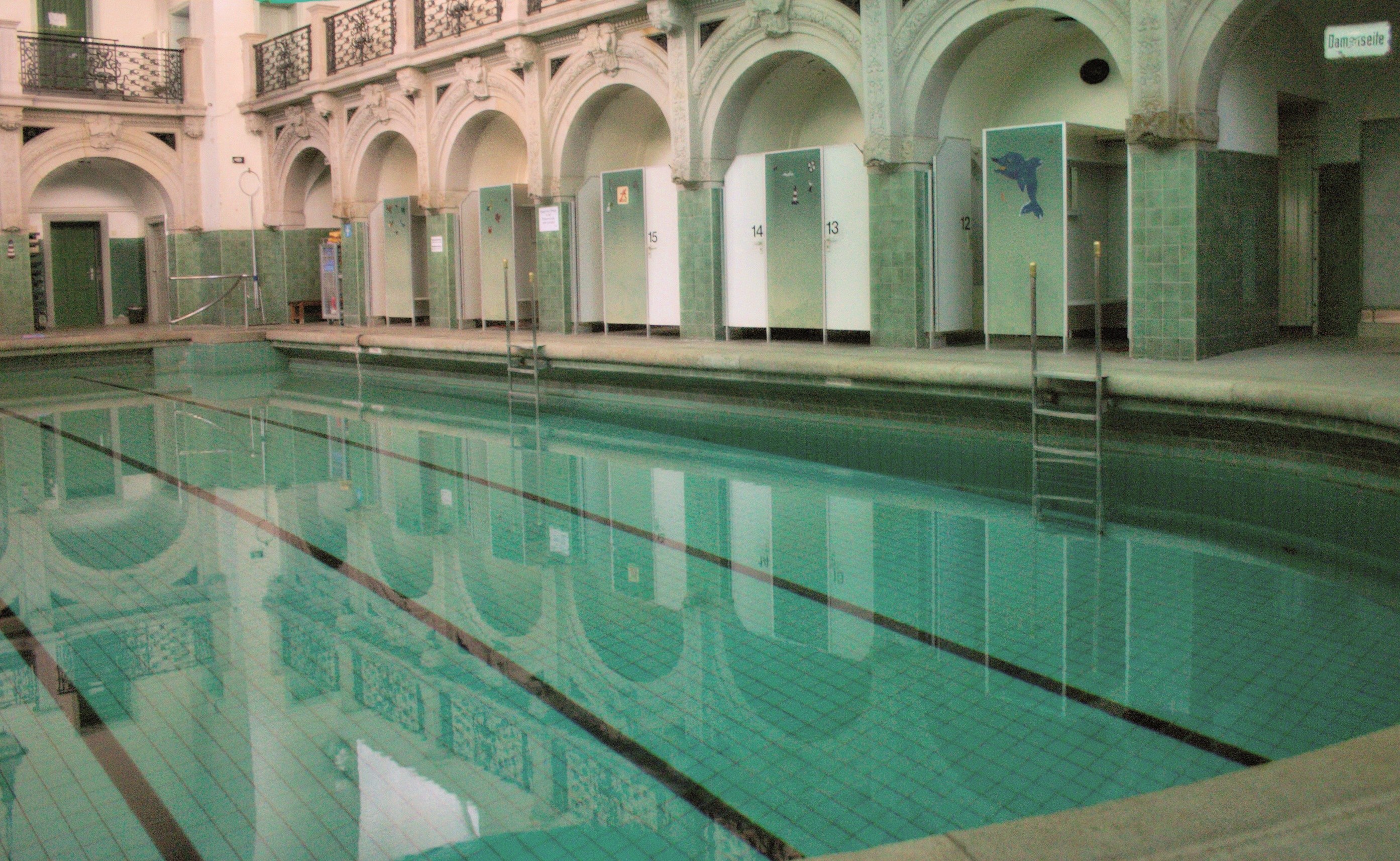 Indoor-Schwimmbad mit kristallklarem Wasser, umgeben von einem gefliesten Boden, in einem großen Gebäude mit Bögen, Säulen, Geländern, Türen und Tafeln mit Text.