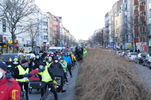 Eine große Gruppe von Menschen mit Masken und Sicherheitswesten auf Fahrrädern auf einer von Bäumen gesäumten Straße mit Gebäuden, Laternenmästen und Texttafeln, begleitet von Fahrzeugen und trockenem Gras, unter einem klaren blauen Himmel.