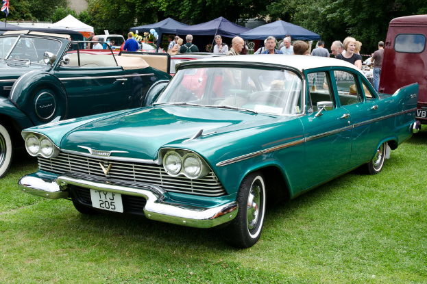 Vintage cars parked on grass with people standing behind them, tents in the background, trees at the top, and a flag in the top left corner.