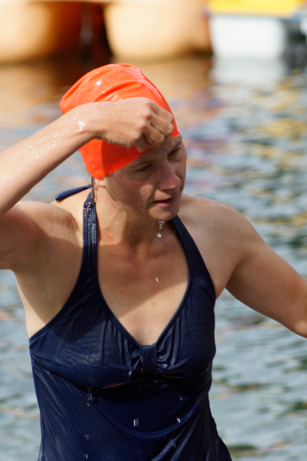 Eine Frau in Badekleidung und Badekappe steht in einem Gewässer, wobei die Beine einer anderen Person im Hintergrund zu sehen sind.