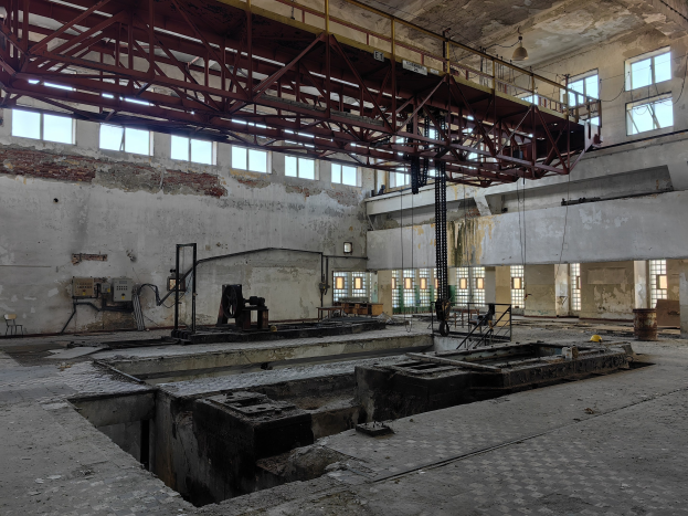 Verlassene Fabrikhalle mit großen Fenstern, Metallstangen und verstreuten industriellen Objekten vor sichtbaren Wänden.