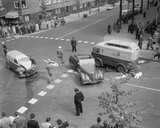 Schwarzes und weißes Foto eines Autounfalls an einer Straßenkreuzung mit einem Van, anderen Fahrzeugen und einer Gruppe von Zuschauern; Gebäude, Bäume, Pfosten und Schilder sind im Hintergrund zu sehen.