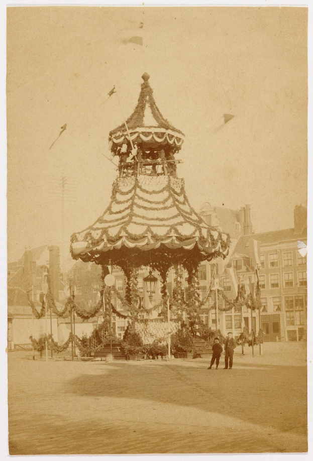 Ein altes Schwarz-Weiß-Foto eines Stadtkarussells, umgeben von Menschen, Pfählen, Lichtern und Gebäuden, geschmückt mit festlichen Weihnachtsdekorationen und mit dem Himmel im Hintergrund.