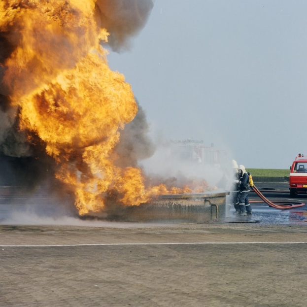 Feuerwehrlöschfahrzeug in Flammen an der Straßenseite mit zwei Helmträgern, die Schläuche halten, einem Fahrzeug im Hintergrund und dem Himmel.