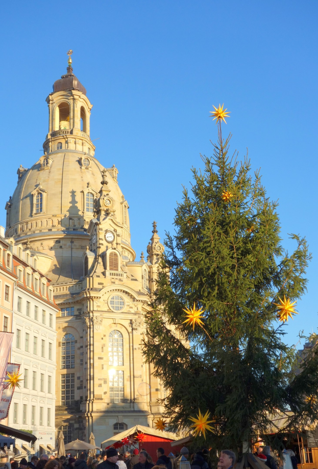 Ein geschäftiger Weihnachtsmarkt in Dresden, Deutschland, mit einem großen geschmückten Weihnachtsbaum vor einer Kirche, umgeben von Menschen, einer Fahne links daneben und einem sichtbaren Himmel im Hintergrund.