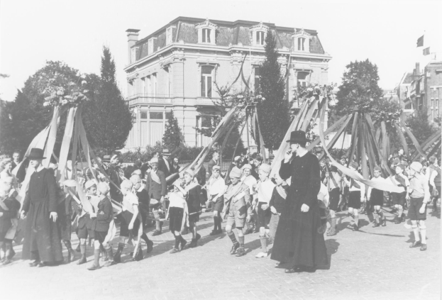 Schwarz-weißes Bild einer Gruppe von Menschen, die eine Straße entlanggehen und Fahnen halten, mit Hüten, Bäumen, Gebäuden und einem Maibaum im Hintergrund.