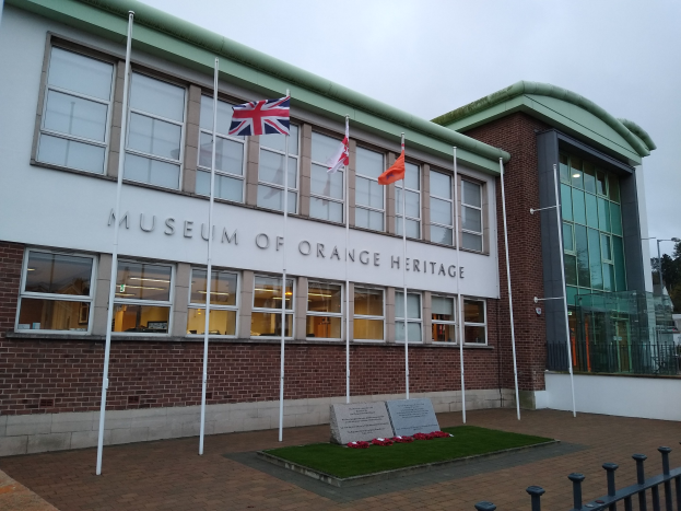 Das Museum of Orange Heritage-Gebäude mit Fenstern und Text, umgeben von Fahnen auf Stangen, Gras, einer Informationswand, einem Zaun, Bäumen, Straßenlaternen und einem bewölkten Himmel.