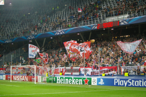 Ein Fussballspiel wird in einem gut beleuchteten Stadion mit einer grossen Zuschauermenge, grünem Rasen, einem Torpfosten mit Bannern und einem Bildschirm in der oberen linken Ecke gespielt.