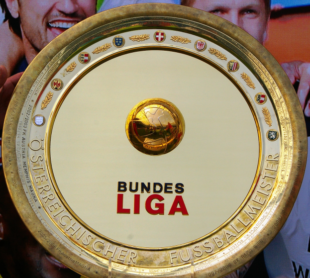 Goldener Schild mit 'Bundesliga'-Text, gehalten von einem Mann, mit einem Banner im Hintergrund, das drei Personen zeigt.