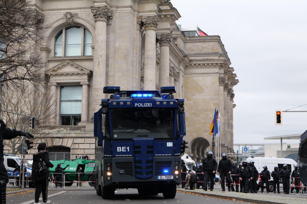 Gruppe von Polizisten vor einem großen Gebäude mit Fenstern, Säulen und Bögen, mit Fahrzeugen auf der Straße und einer Person mit einer Kamera auf der linken Seite.