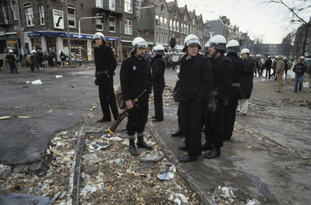 Eine Gruppe von Polizisten in Helmen und mit Waffen in der Hand steht auf der Seite einer Straße mit Gebäuden, Schildern, Laternen, Bäumen und einem klaren Himmel im Hintergrund.