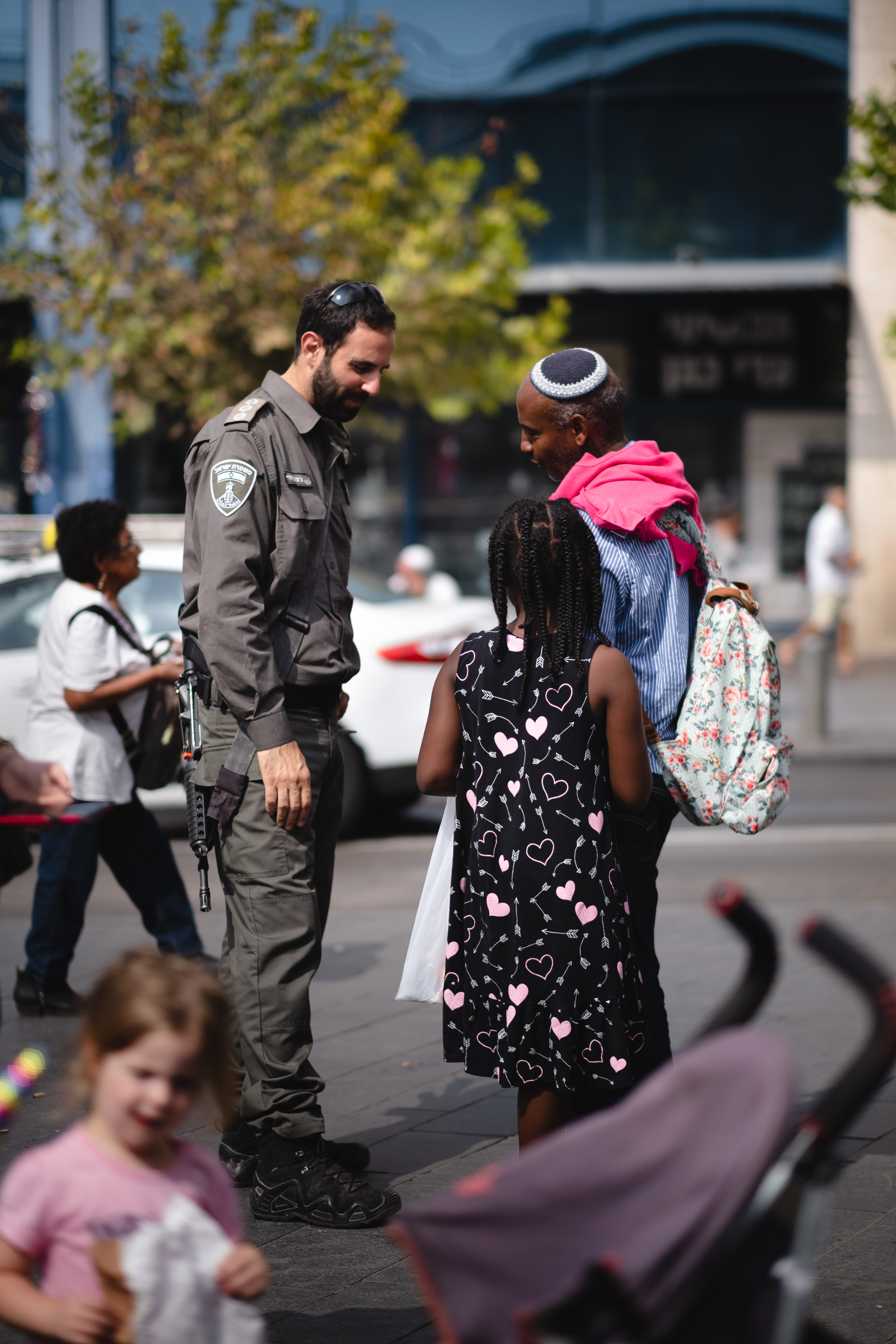 Ein Polizeibeamter spricht zu einer Gruppe von Menschen auf der Straße, mit einem Kinderwagen im Vordergrund und Fahrzeugen, Bäumen und einem Gebäude im Hintergrund.