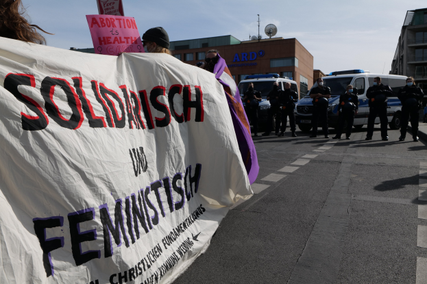 Eine Gruppe von Menschen marschiert mit einem "Solidarität und Feminismus"-Schild die Straße entlang, mit parkenden Fahrzeugen, Gebäuden, einer Schüsselantenne und einem klaren blauen Himmel im Hintergrund.