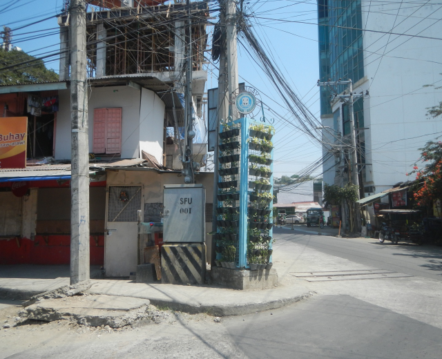 Stadtstraßenszene mit Gebäuden, Strommasten, Drähten, Fahrzeugen, Bäumen und einem Himmel als Hintergrund, mit einem Gebäude mit Drähten an der Seite im Vordergrund.