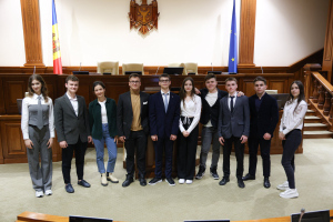 Gruppe von Menschen, die vor einem Gerichtssaal mit Monitoren und Mikrofonen auf Tischen, einer hölzernen Plattform mit Stühlen, Flaggen und einem an der Wand montierten Objekt im Hintergrund und einer Treppe mit Geländern auf der rechten Seite posieren.