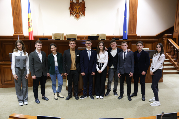 Gruppe von Menschen, die vor einem Gerichtssaal mit Monitoren und Mikrofonen auf Tischen, einer hölzernen Plattform mit Stühlen, Flaggen und einem an der Wand montierten Objekt im Hintergrund und einer Treppe mit Geländern auf der rechten Seite posieren.