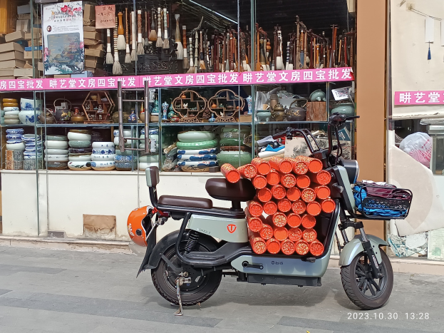 Ein Motorroller mit einem Korb steht auf der Straße vor einem Laden mit einer Glaswand, auf der verschiedene Gegenstände zu sehen sind, und eine Tafel mit Text ist auf der Ladenfront sichtbar.