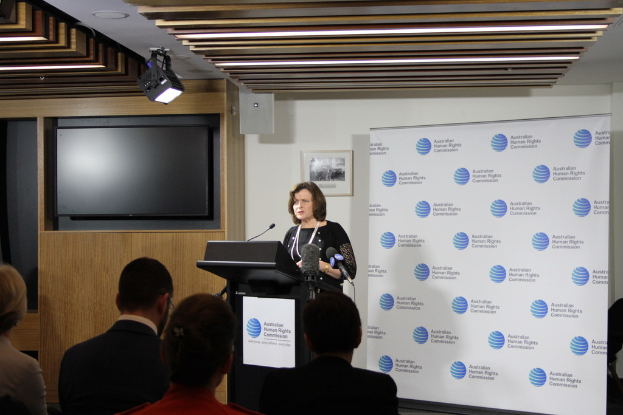 Eine Frau steht auf einer Bühne und spricht in ein Mikrofon, vor ihr sitzen Zuschauer, hinter ihr eine Wand mit einem Banner, ein Fernseher an der Holzwand und Lampen an der Decke.