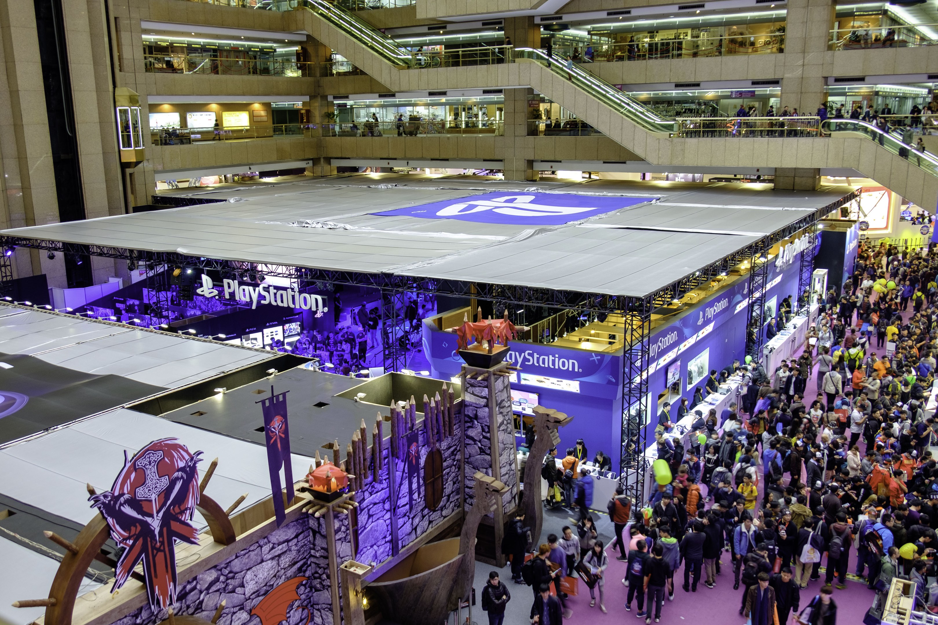 Eine große Menschenmenge in einer großen Indoor-Venue mit Rolltreppen, Geländern, beleuchteten Schildern, Bannern und anderen Anzeigen, die als die größte Gaming-Convention der Welt identifiziert wird.