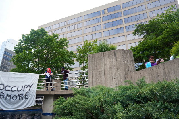 Menschen auf einer Brücke mit einem Banner und einem Fahrrad, unter der sich Wasser befindet, mit Bäumen, Gebäuden und Himmel im Hintergrund.