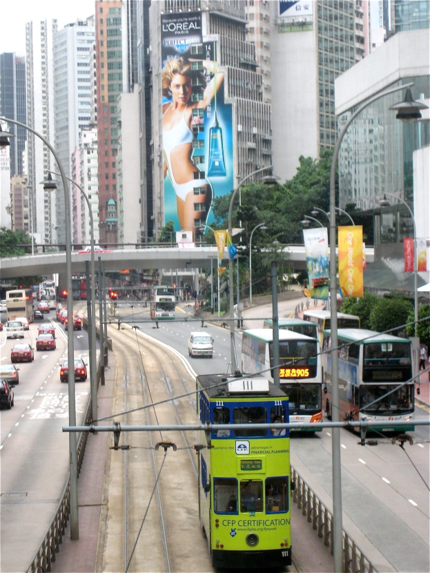 Eine belebte Stadtstraße mit Doppeldeckerbussen, Autos und anderen Fahrzeugen, mit Polen, Laternen, Schildern, Bäumen, einer Brücke, hohen Gebäuden und einem klaren blauen Himmel, mit Text am unteren Rand.