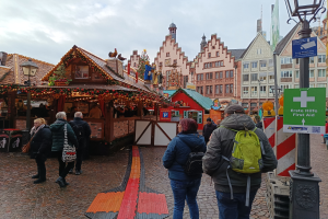 Eine Gruppe von Menschen geht eine Kopfsteinpflasterstraße entlang neben einem Weihnachtsmarkt in Nürnberg, Deutschland, mit Taschen, Laternenmasten und Texttafeln tragend, und Gebäuden mit Fenstern im Hintergrund unter einem bewölkten Himmel.