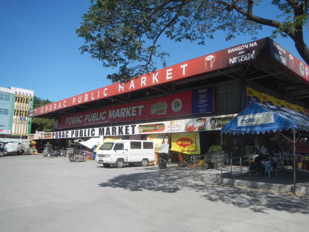 Ein ├Âffentlicher Markt mit einem vor ihm geparkten Van, umgeben von Geb├Ąuden, Gesch├Ąften, Namensschildern, Kraftfahrzeugen auf der Stra├če, Personen unter einem Zelt sitzend, Gel├Ąndern, B├Ąumen und einem klaren blauen Himmel.