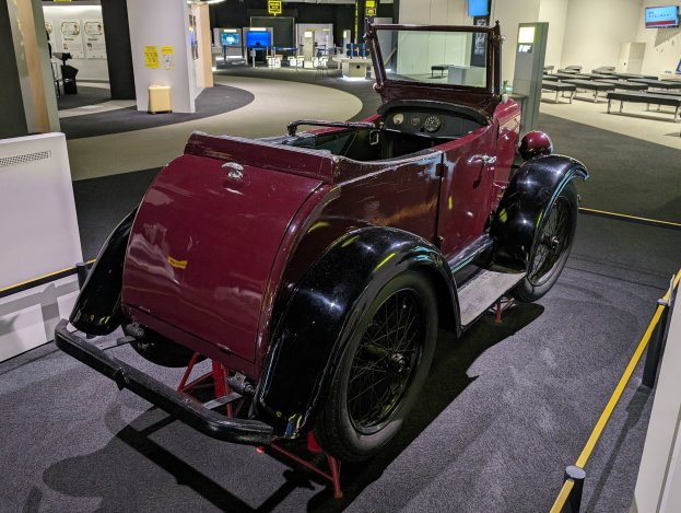 Alter roter Wagen in einem Museum mit Absperrpollern, Bändern, Tischen, Stühlen, Bildschirmen, Säulen, Texttafeln und Beleuchtung ausgestellt.