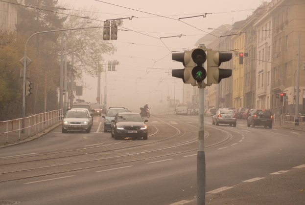 Eine städtische Straße voller Verkehr an einem nebligen Tag, mit Fahrzeugen, einem Radfahrer, Verkehrsampeln, Laternen, Schildern, Geländern, Bäumen, Gebäuden mit Fenstern und einem Himmel im Hintergrund.