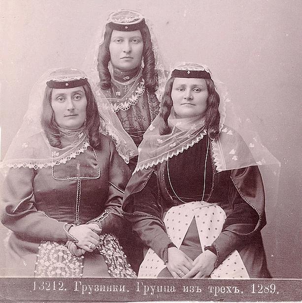 Drei Frauen in traditionellen russischen Hochzeitskleidern mit farbenfroher Stickerei und Perlen, die vor einer einfarbigen weißen Wand in einem Schwarz-Weiß-Foto posieren.